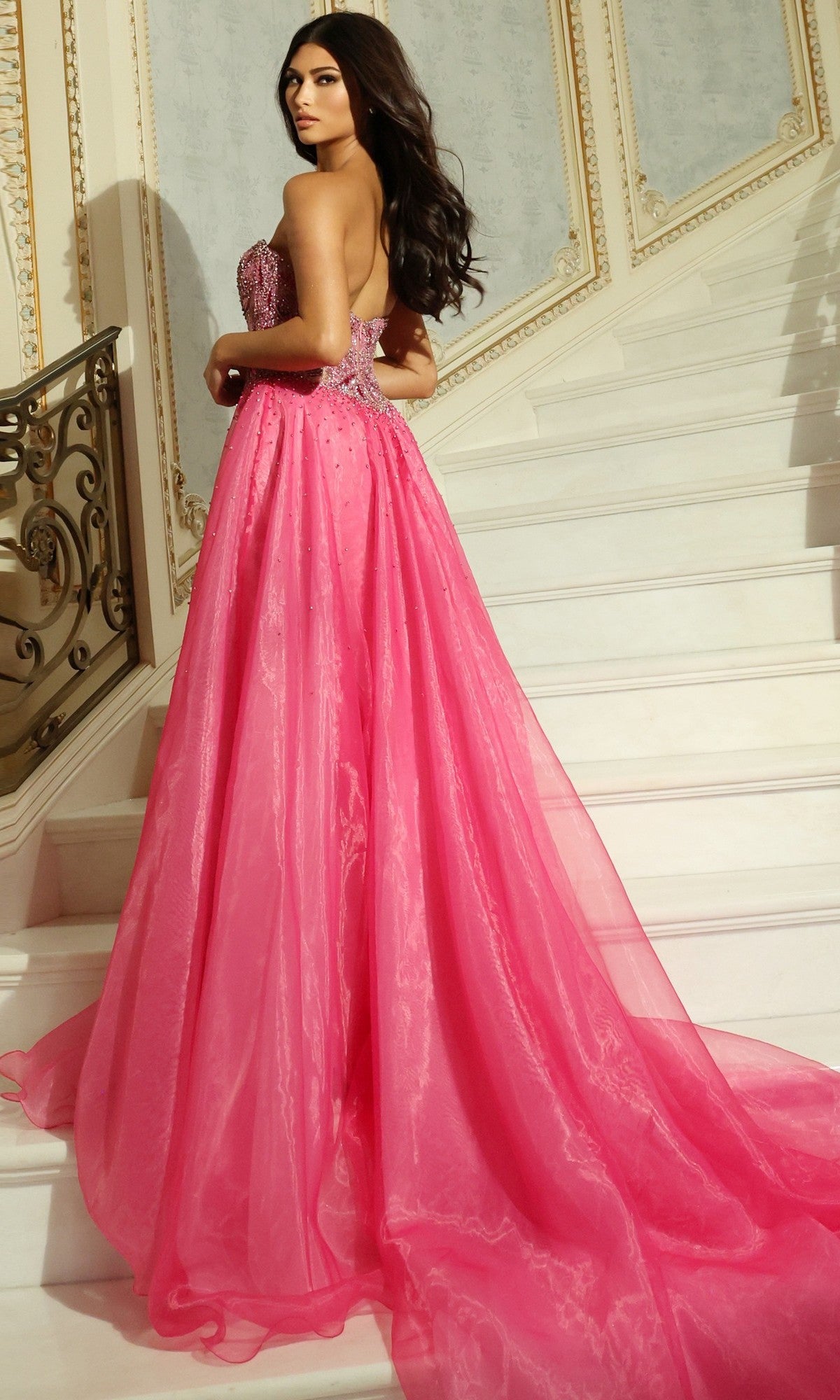 A woman wearing the Jovani 48906 long prom dress with a strapless beaded corset bodice and an A-line organza skirt stands on a white staircase with ornate railings and decorative walls.